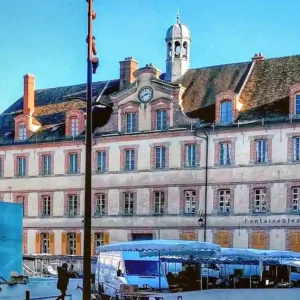 Place de la République à Fontainebleau pendant un jour de marché. On y voit des étals de marché couverts de bâches bleues et blanches, avec quelques visiteurs et vendeurs. Au fond, on aperçoit des bâtiments historiques aux façades de couleur crème, avec des volets de fenêtre marron et des toits en ardoise. Le bâtiment principal, probablement un lieu notable, présente des détails architecturaux comme des fenêtres en arc et un petit clocher. La scène est baignée d'une lumière vive, suggérant qu'il s'agit d'une journée ensoleillée.